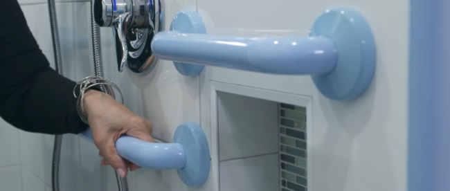 The image shows a close-up of a person's hand gripping a blue grab bar in a bathroom setting. The grab bar is mounted on a white tiled wall, next to a silver shower unit that has a chrome finish. There is a shower hose attached to the unit, and below the grab bar, there is a recessed niche in the wall that contains a soap dish with a bar of soap.